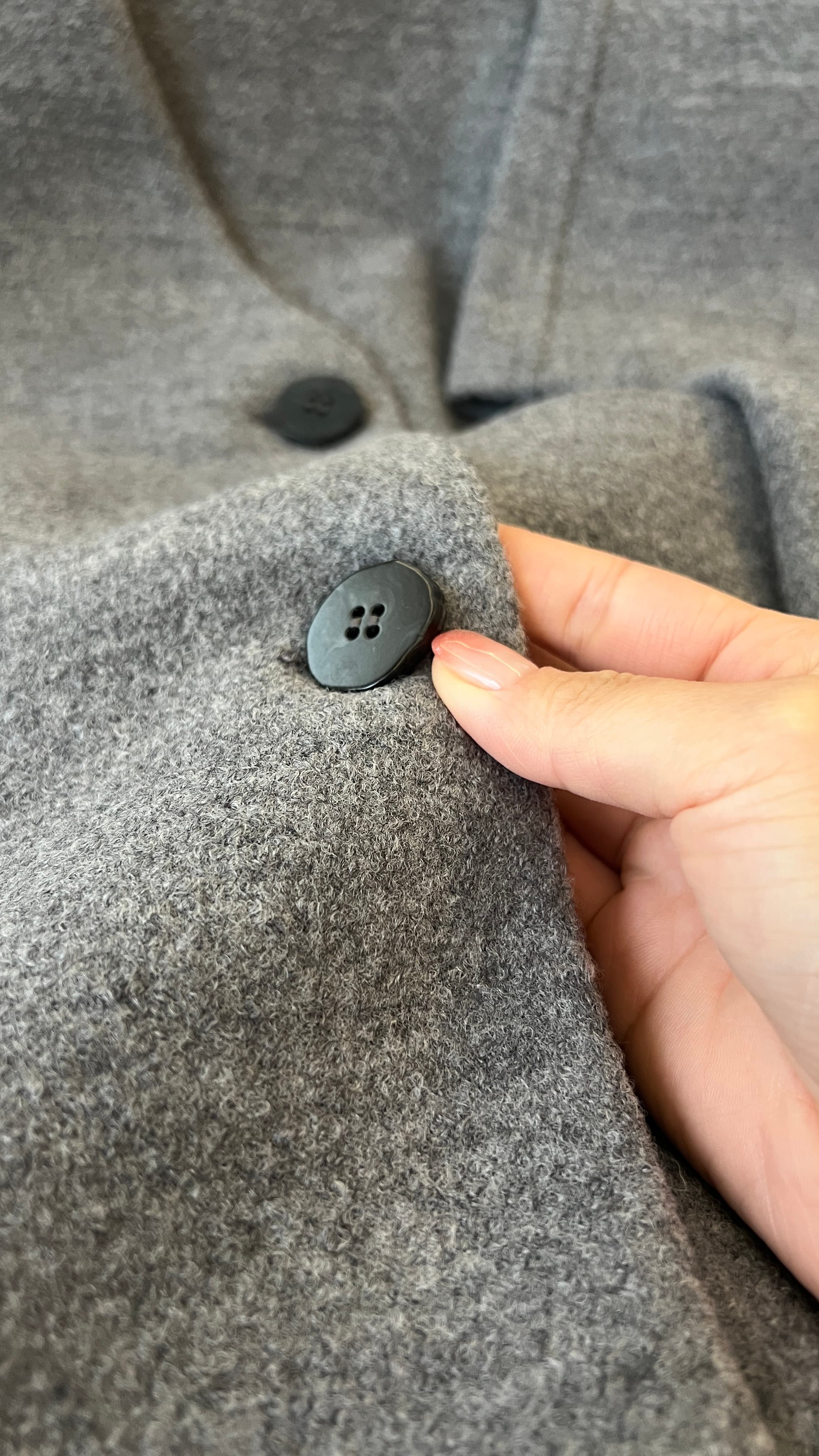A hand pressing a matte black button into soft grey boiled wool — a tactile QC check before a brand garment ships